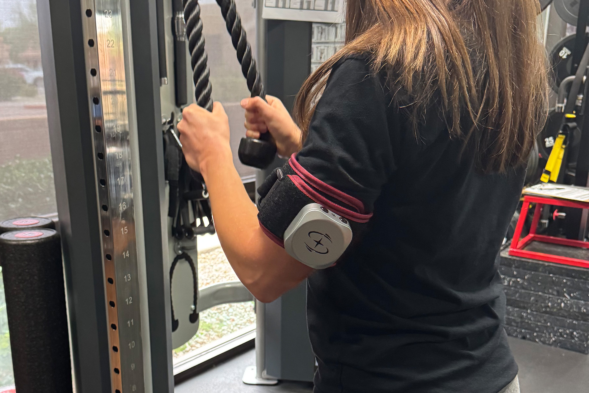 A woman is wearing a BFR cuff while performing low weight rope pull downs at Synergy Physical Therapy