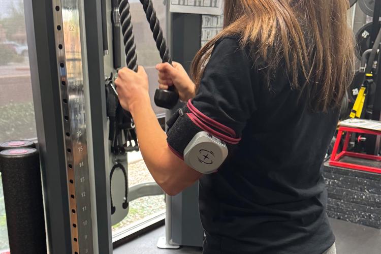 A woman is wearing a BFR cuff while performing low weight rope pull downs at Synergy Physical Therapy